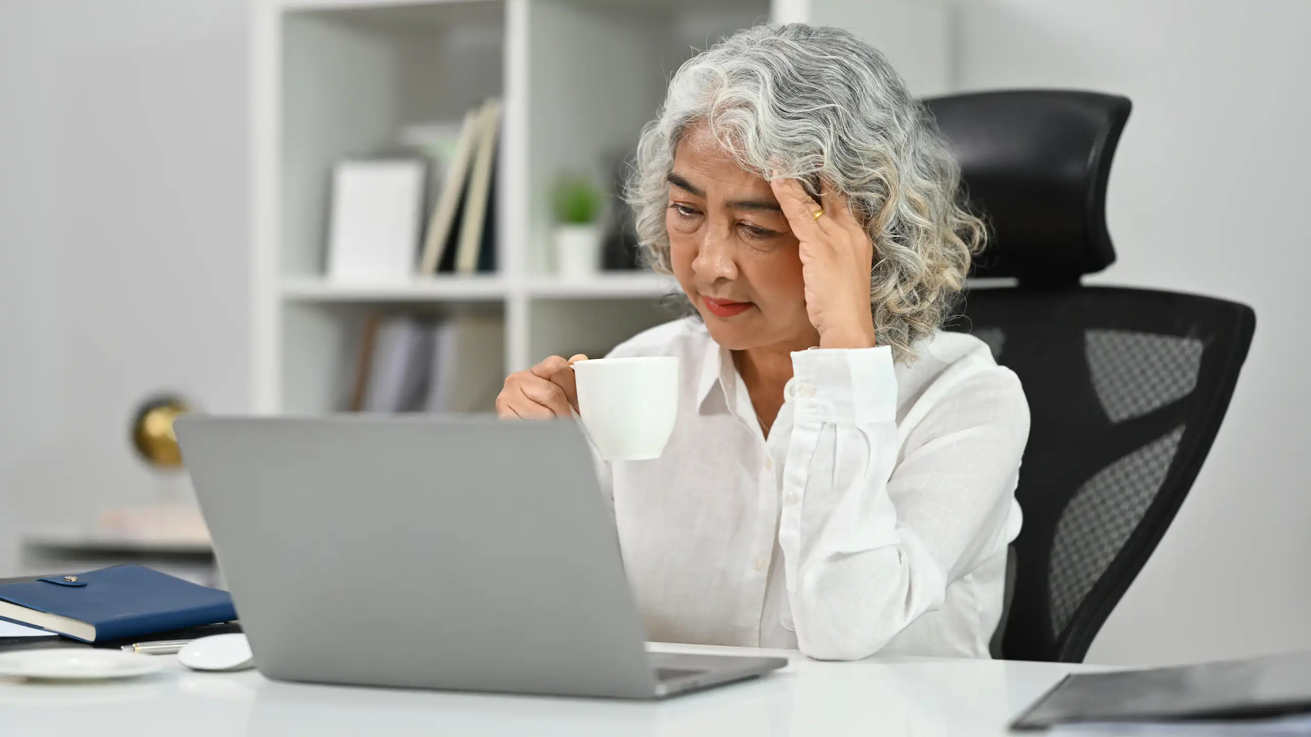 Une femme de plus de 50 ans travaille devant son ordinateur, en se tenant la tête avec une main.