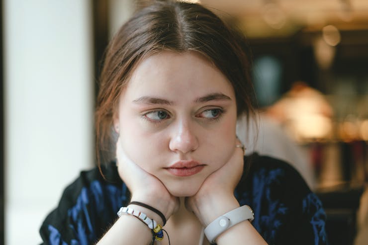 A young girl sits, chin in hands, looking sideways with a serious expression.