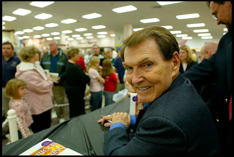 A brown-haired main in a suit grins as he sits at a table in front of a large crowd inside.