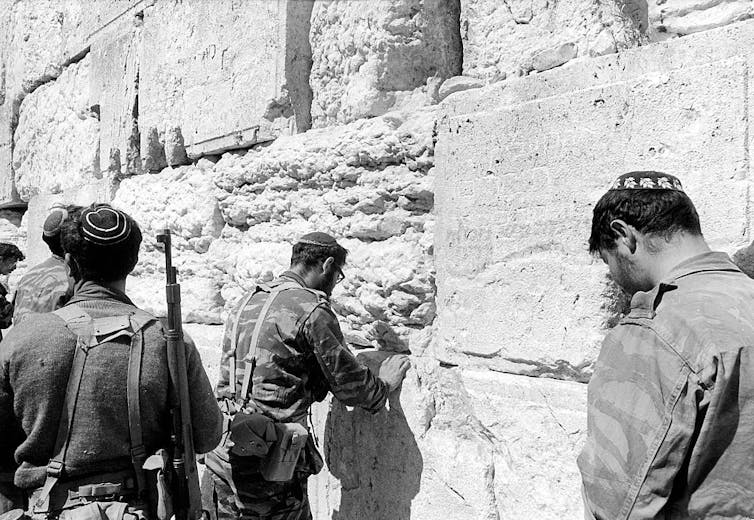 A black and white photo shows the backs of three men in military uniforms facing a wall of large, rough stone blocks.