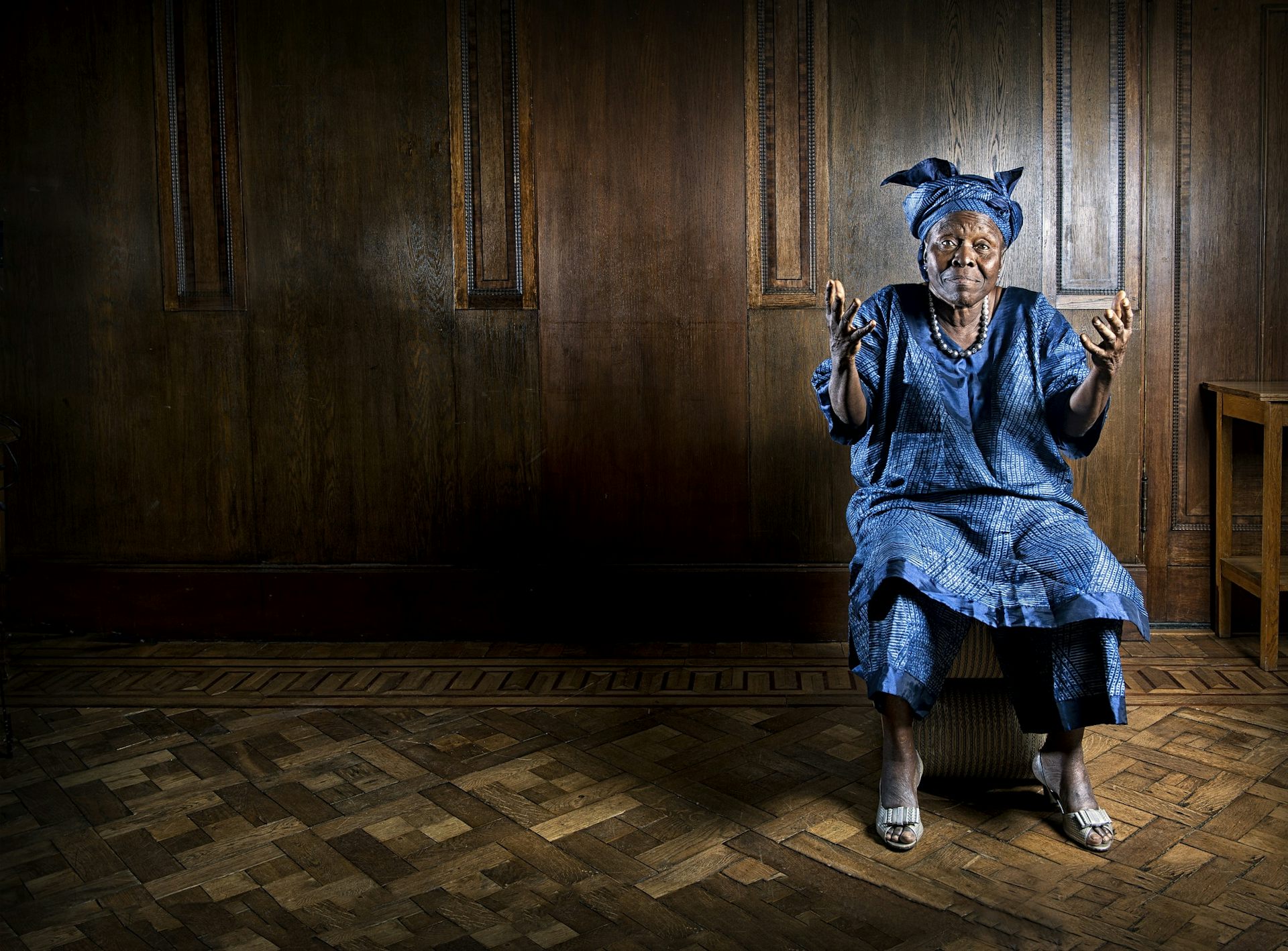 An elder African woman in blue traditional attire poses for a portrait, her hands raised, her expression defiant but bemused