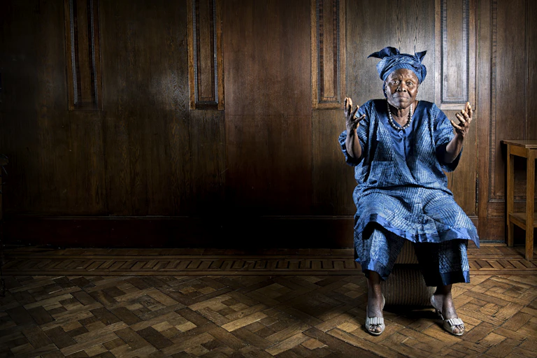 An elder African woman in blue traditional attire poses for a portrait, her hands raised, her expression defiant but bemused