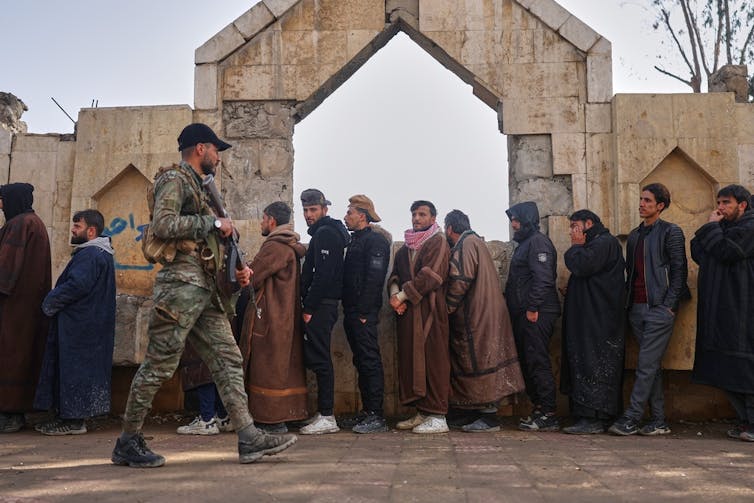 A soldier patrols while a group of men wait in line.