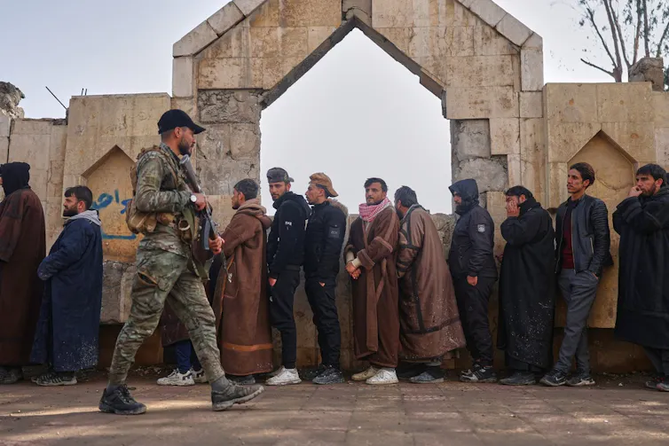 A soldier patrols while a group of men wait in line.