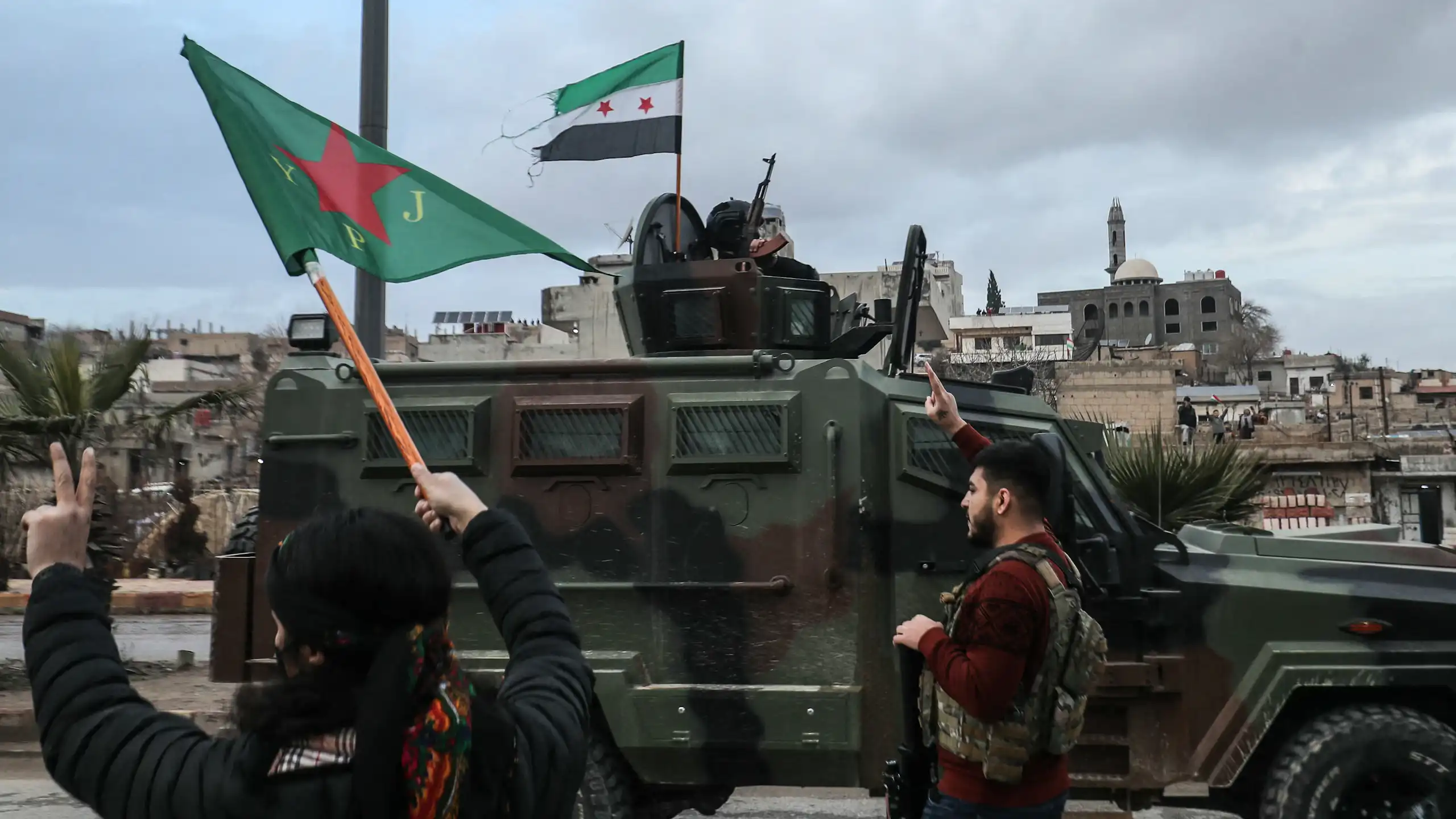 Two people gesture toward an approaching military vehicle.