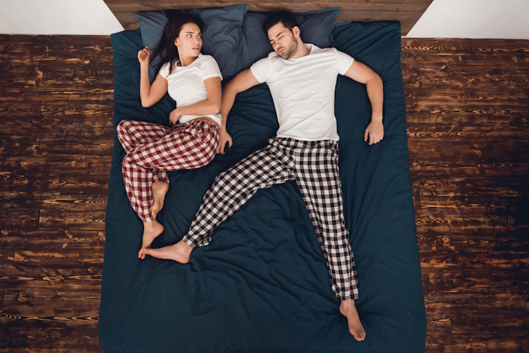 Sleeping man takes up most of a double bed while woman lies awake and stares at her partner in irritation