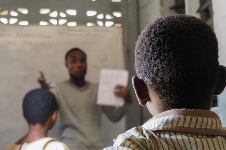 A man at the front of a classroom talking to children and holding up a piece of white paper, with two learners in the frame facing forward
