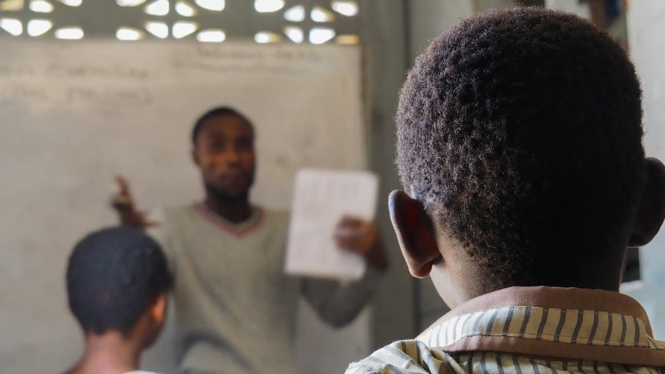 A man at the front of a classroom talking to children and holding up a piece of white paper, with two learners in the frame facing forward