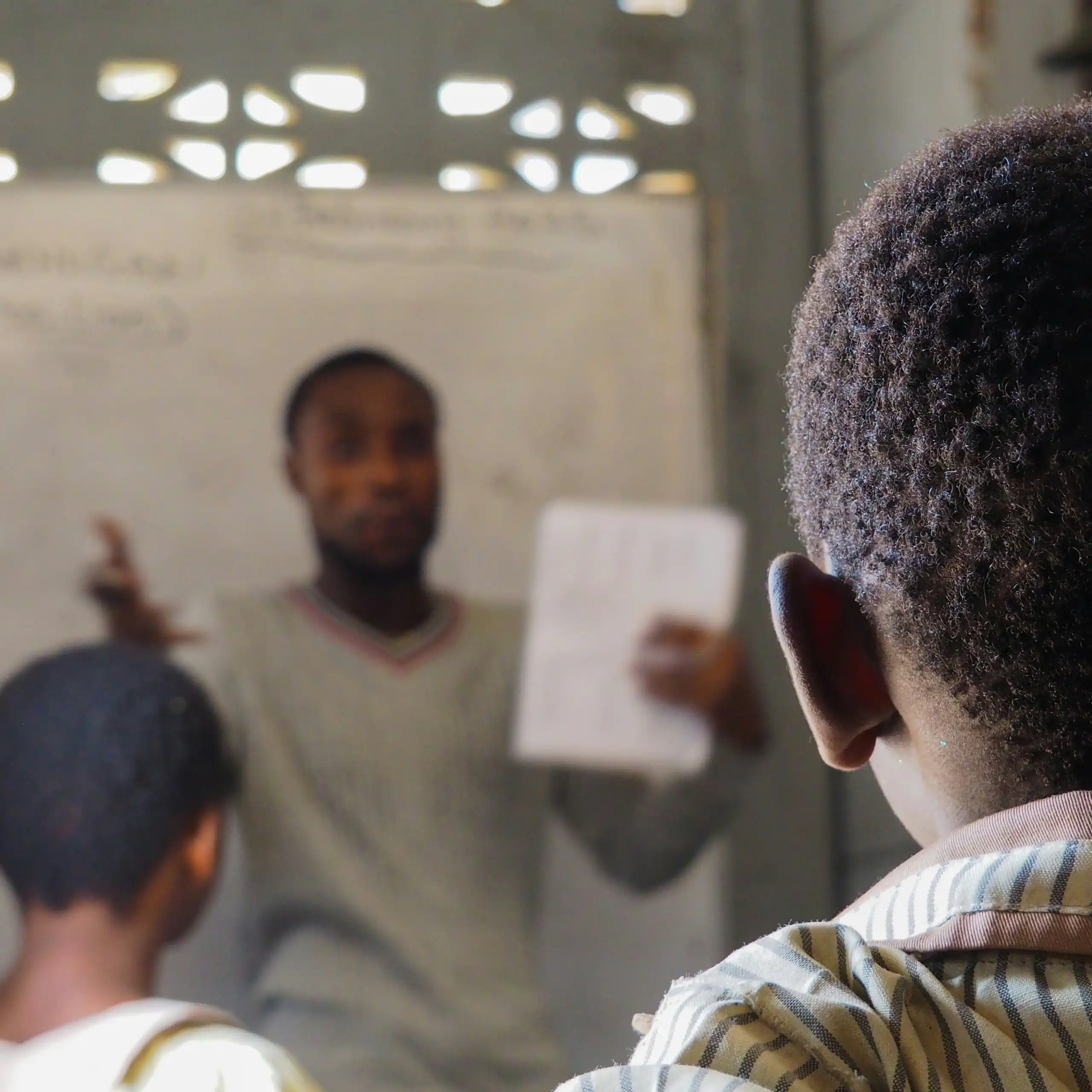 A man at the front of a classroom talking to children and holding up a piece of white paper, with two learners in the frame facing forward