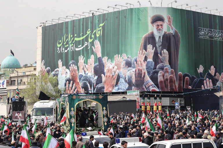 A large crowd of people stand in a street with red, white and green flags and a large billboard that shows a man wearing a robe, waving to people holding their arms up to him.