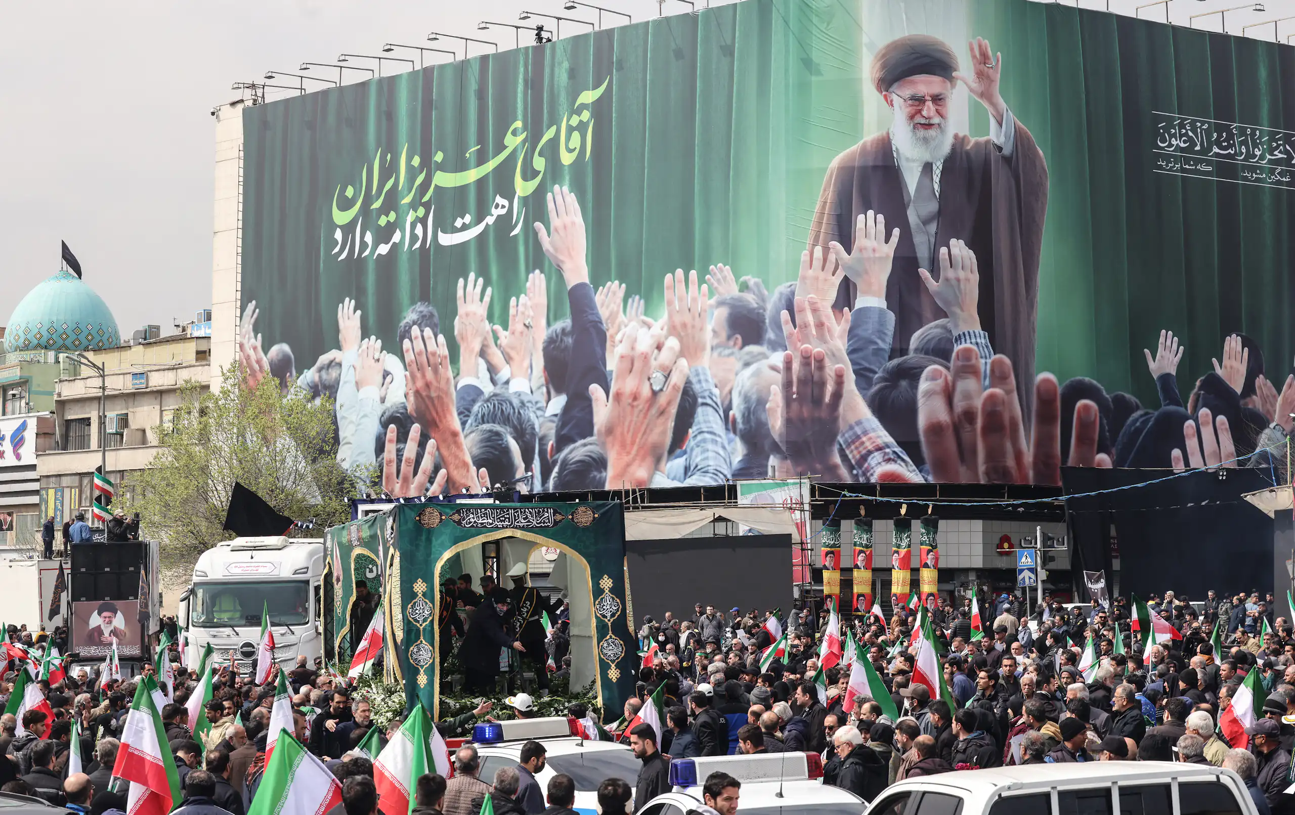 A large crowd of people stand in a street with red, white and green flags and a large billboard that shows a man wearing a robe, waving to people holding their arms up to him.