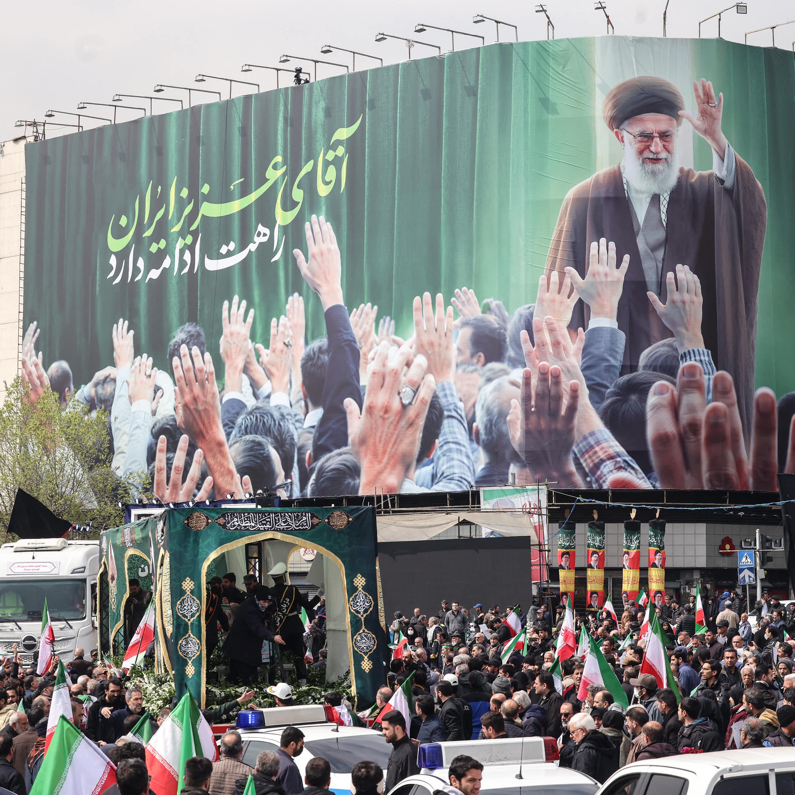 A large crowd of people stand in a street with red, white and green flags and a large billboard that shows a man wearing a robe, waving to people holding their arms up to him.