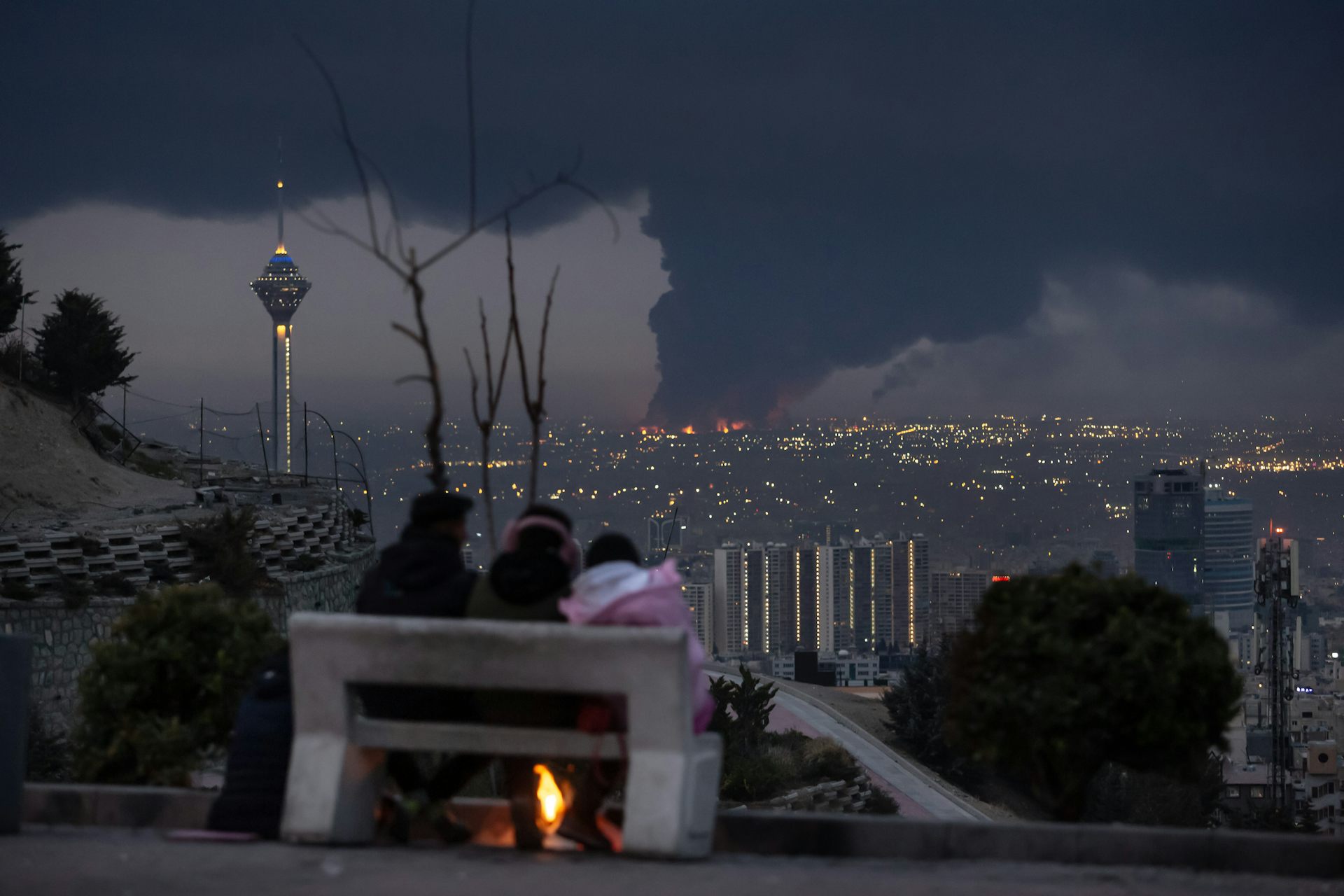 Tres personas sentadas en un banco contemplan la ciudad de la que se eleva un humo oscuro.