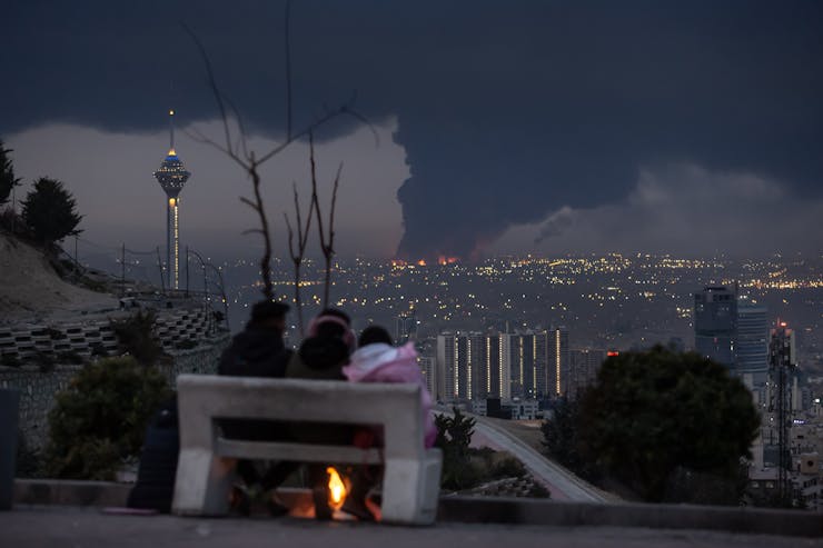 Three people sit on a bench and look over a city that has dark smoke rising from it.