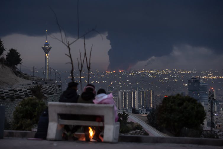 Tres personas sentadas en un banco contemplan la ciudad de la que se eleva un humo oscuro.