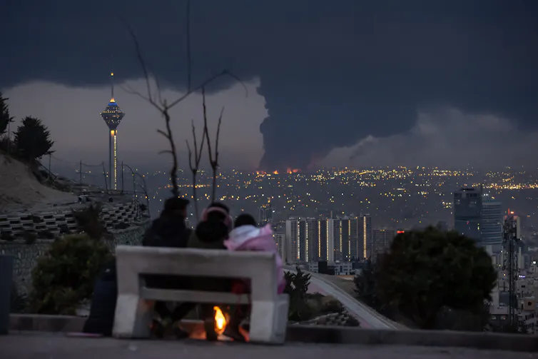 Three people sit on a bench and look over a city that has dark smoke rising from it.
