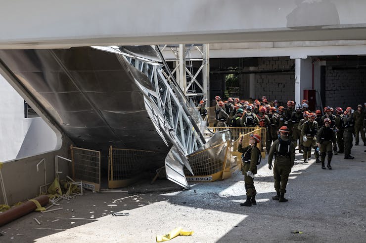 A group of people wearing uniforms gather near a large steel, collapsed structure that appears to be part of a bridge