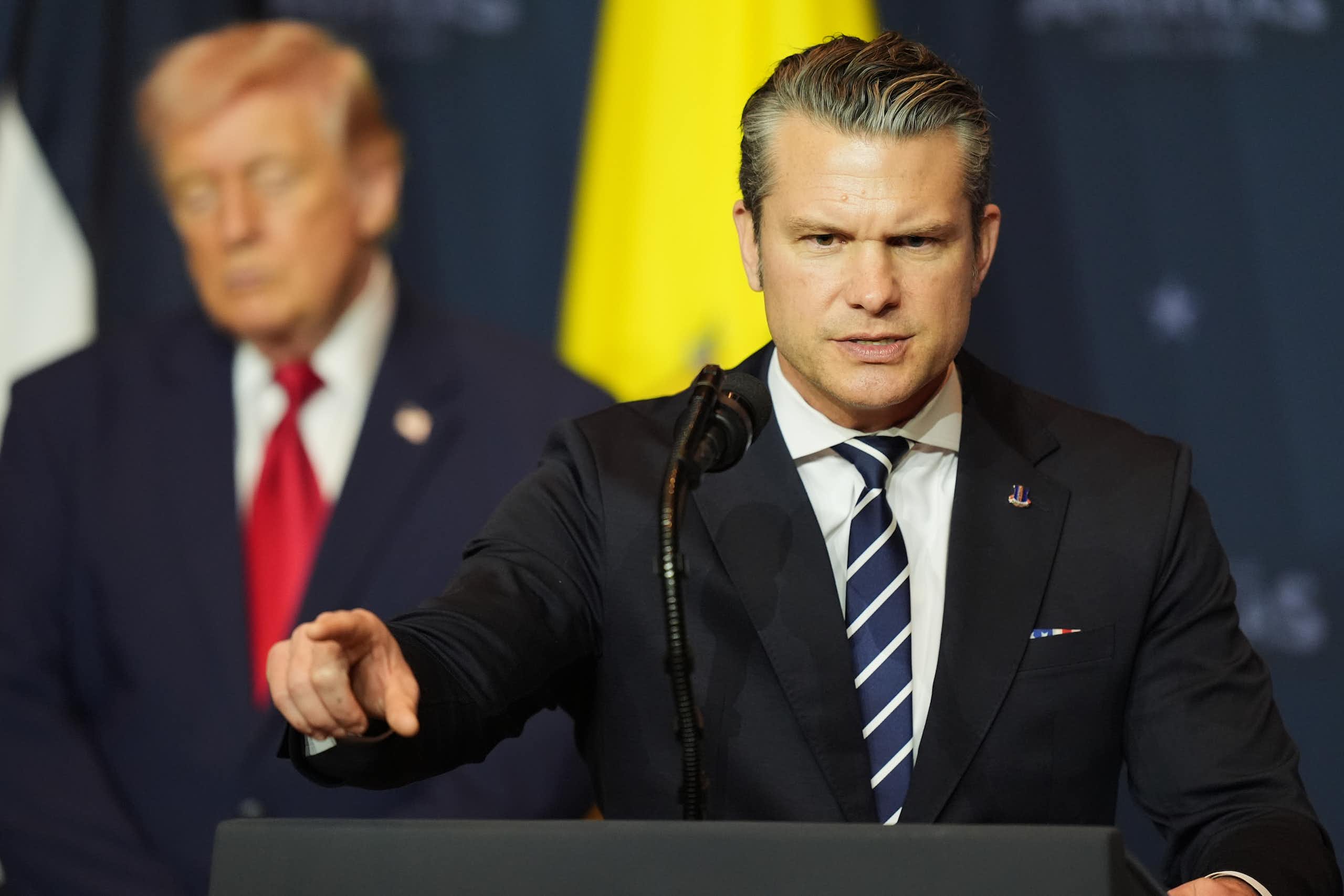an angry Pete Hegseth at podium, Donald Trump behind him