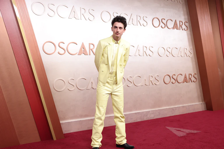 Actor Timothee Chalamet stands on the red carpet at an Oscars ceremony, dressed in a pale yellow suit and with his hands behind his back.