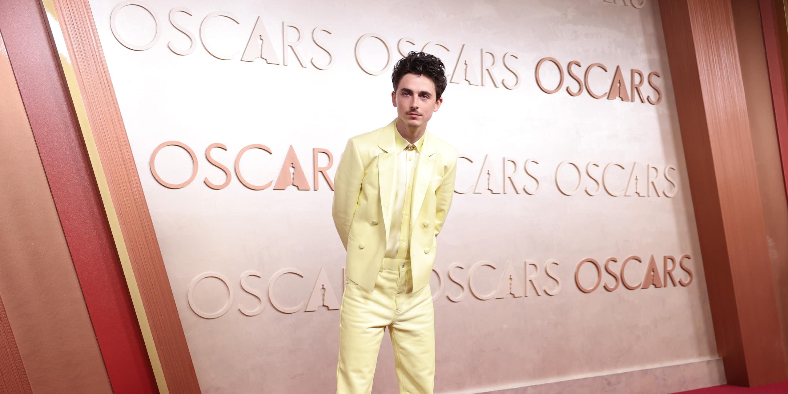 Actor Timothee Chalamet stands on the red carpet at an Oscars ceremony, dressed in a pale yellow suit and with his hands behind his back.