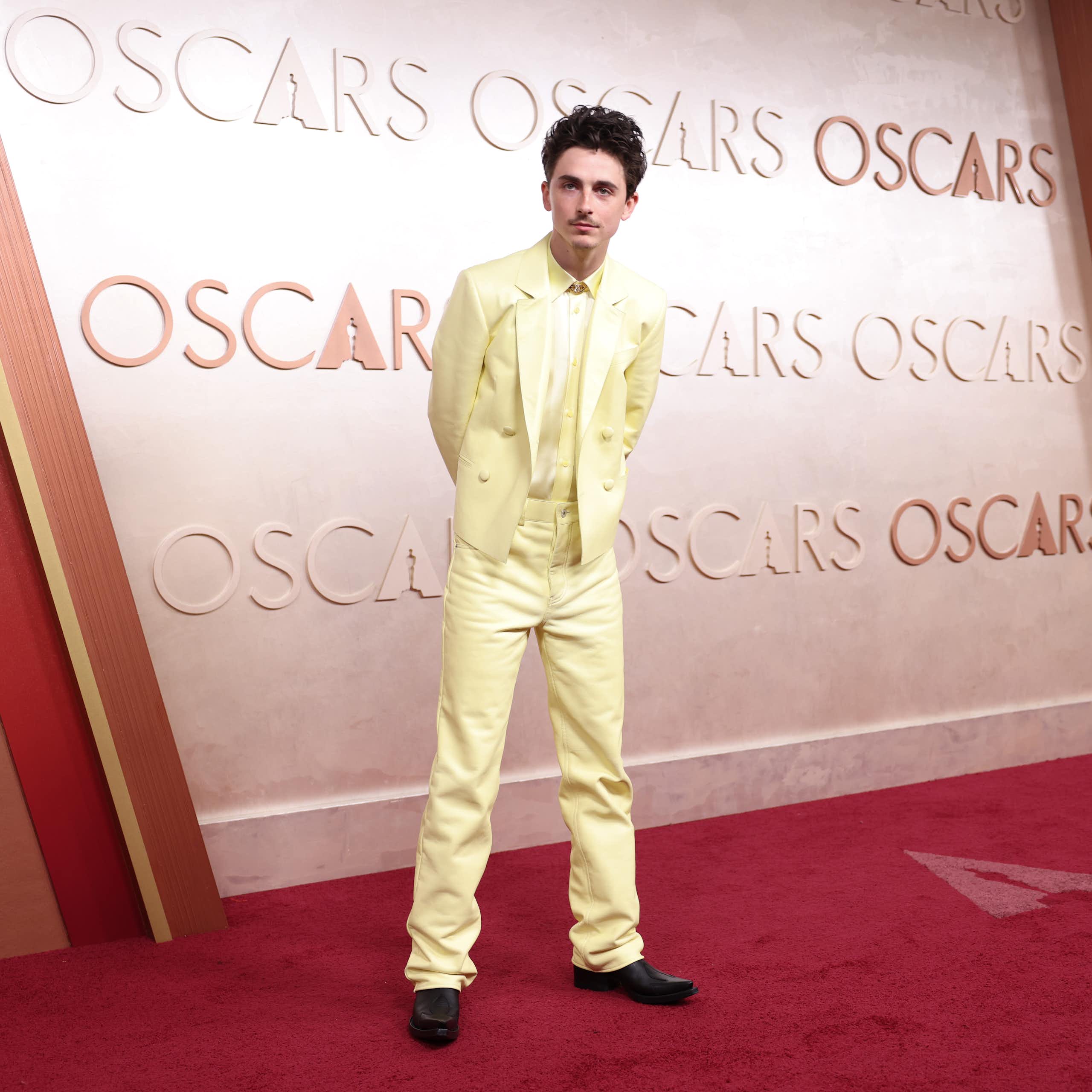 Actor Timothee Chalamet stands on the red carpet at an Oscars ceremony, dressed in a pale yellow suit and with his hands behind his back.