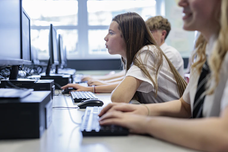 High school students work at row of desk top computers. One student leans forward to peer at her screen.