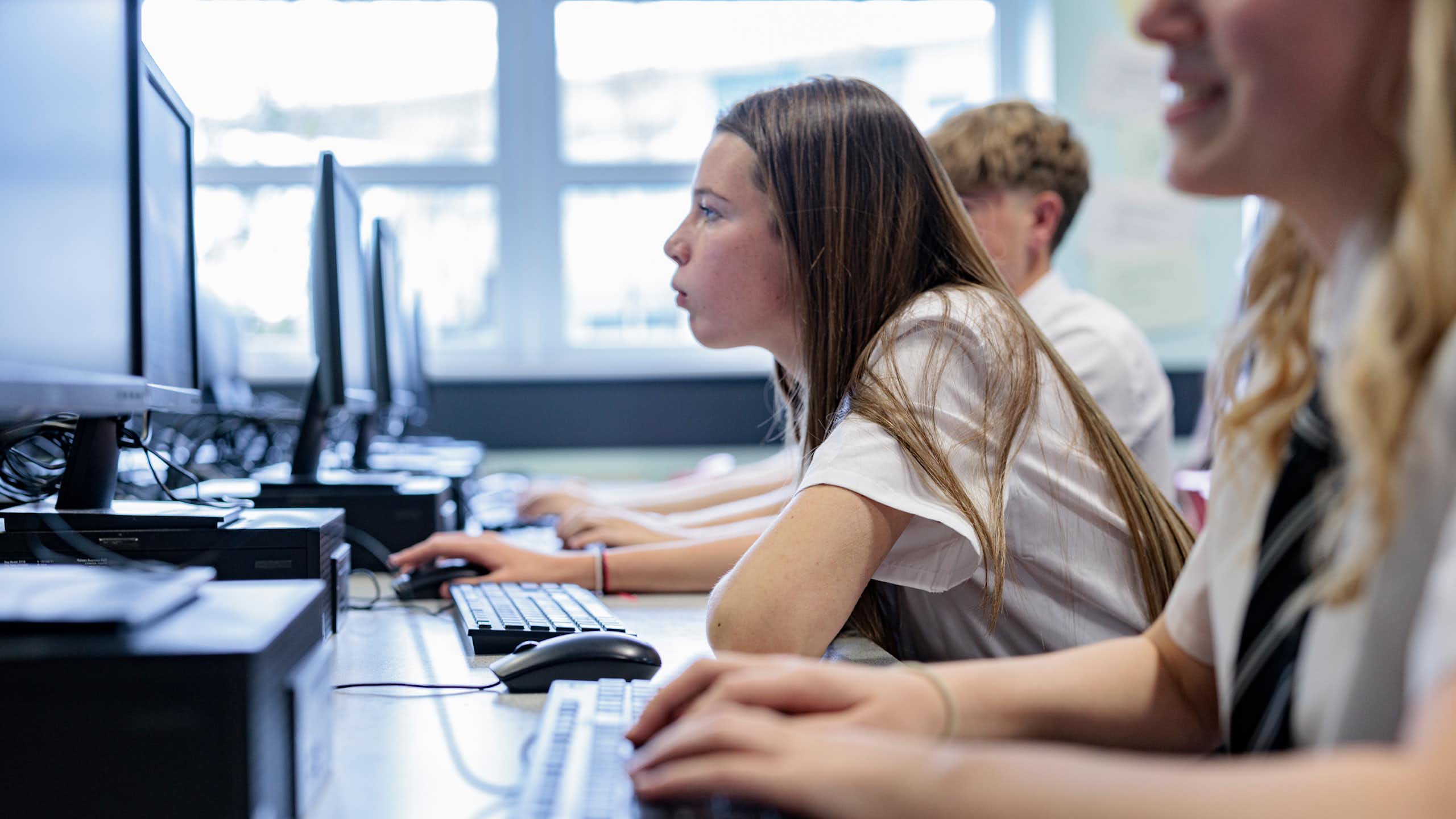 High school students work at row of desk top computers. One student leans forward to peer at her screen.