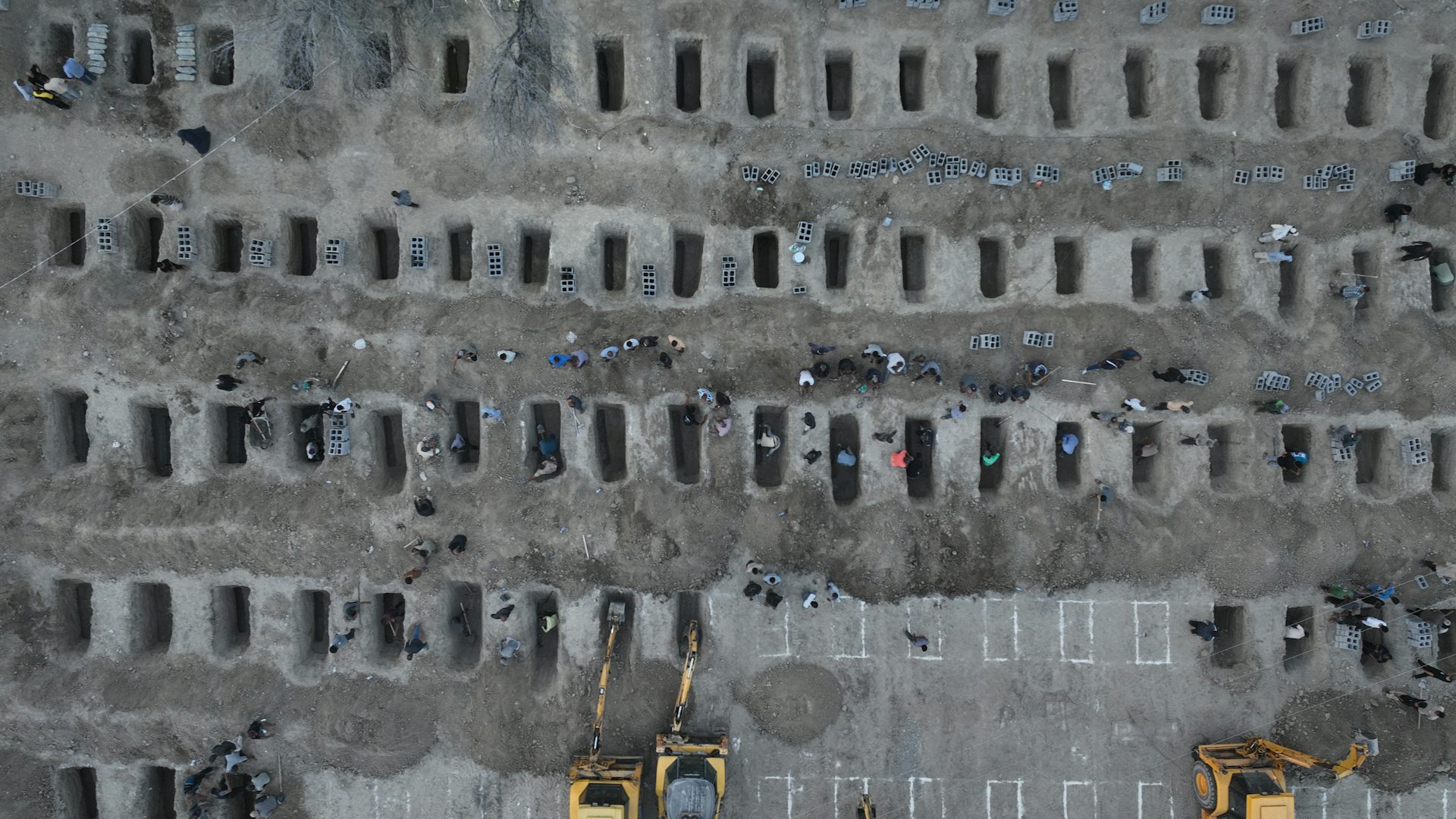 An aerial view shows a mass grave site, with some people looking over the rows of empty graves.