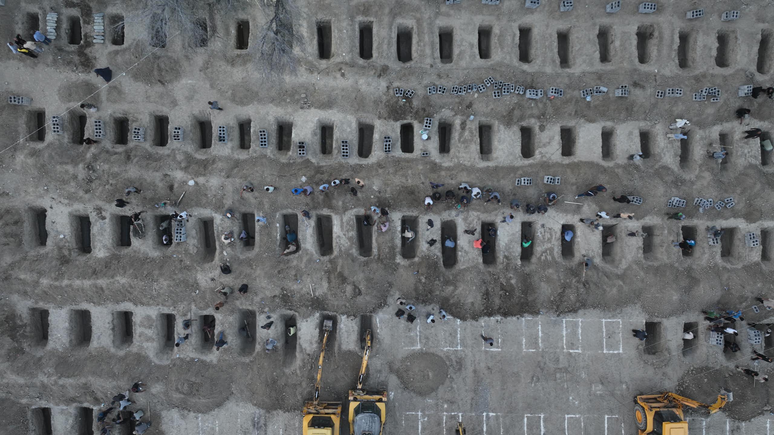 An aerial view shows a mass grave site, with some people looking over the rows of empty graves.