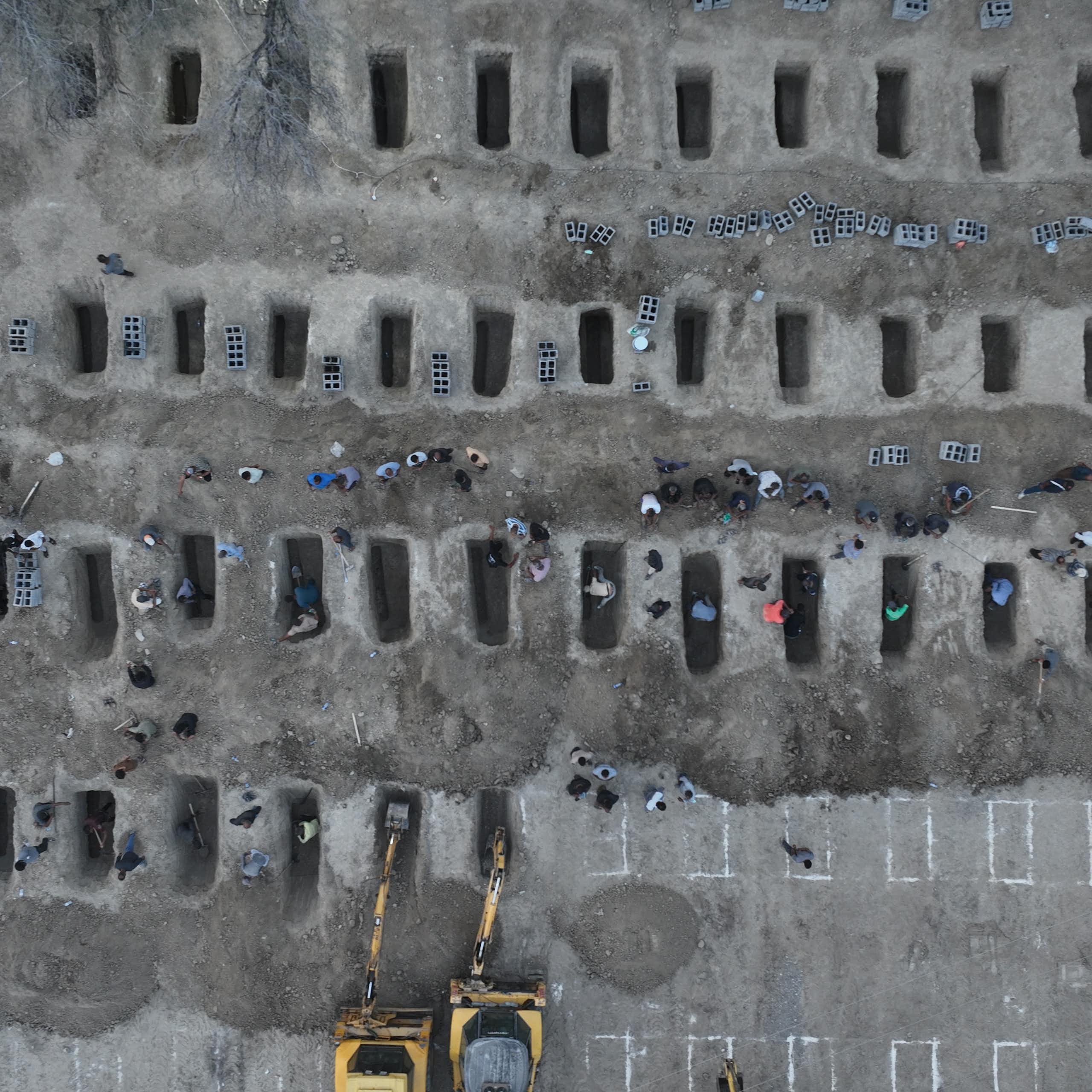 An aerial view shows a mass grave site, with some people looking over the rows of empty graves.