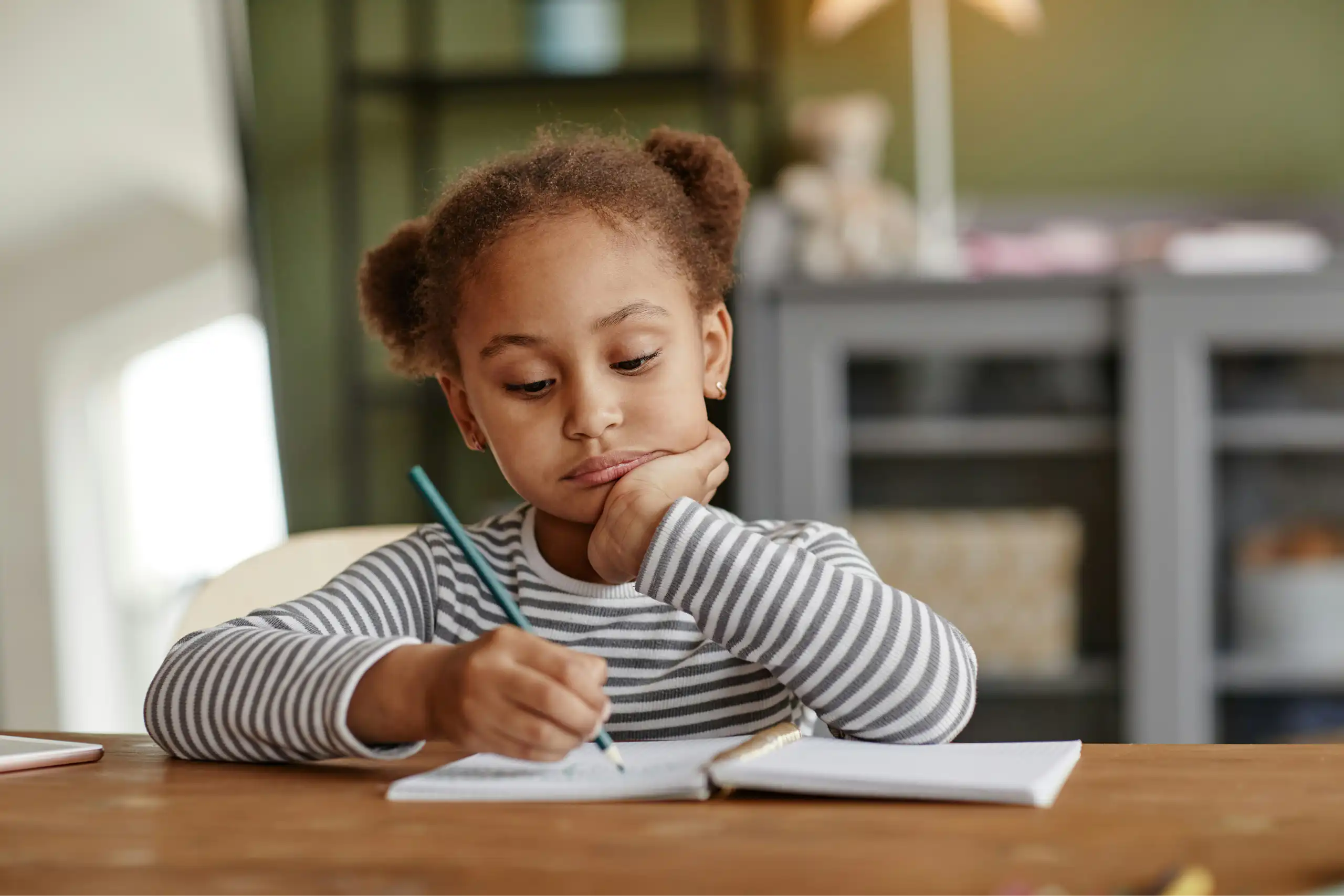 A girl in a striped tee sitting at a table with a notebook, resting her chin in her hand