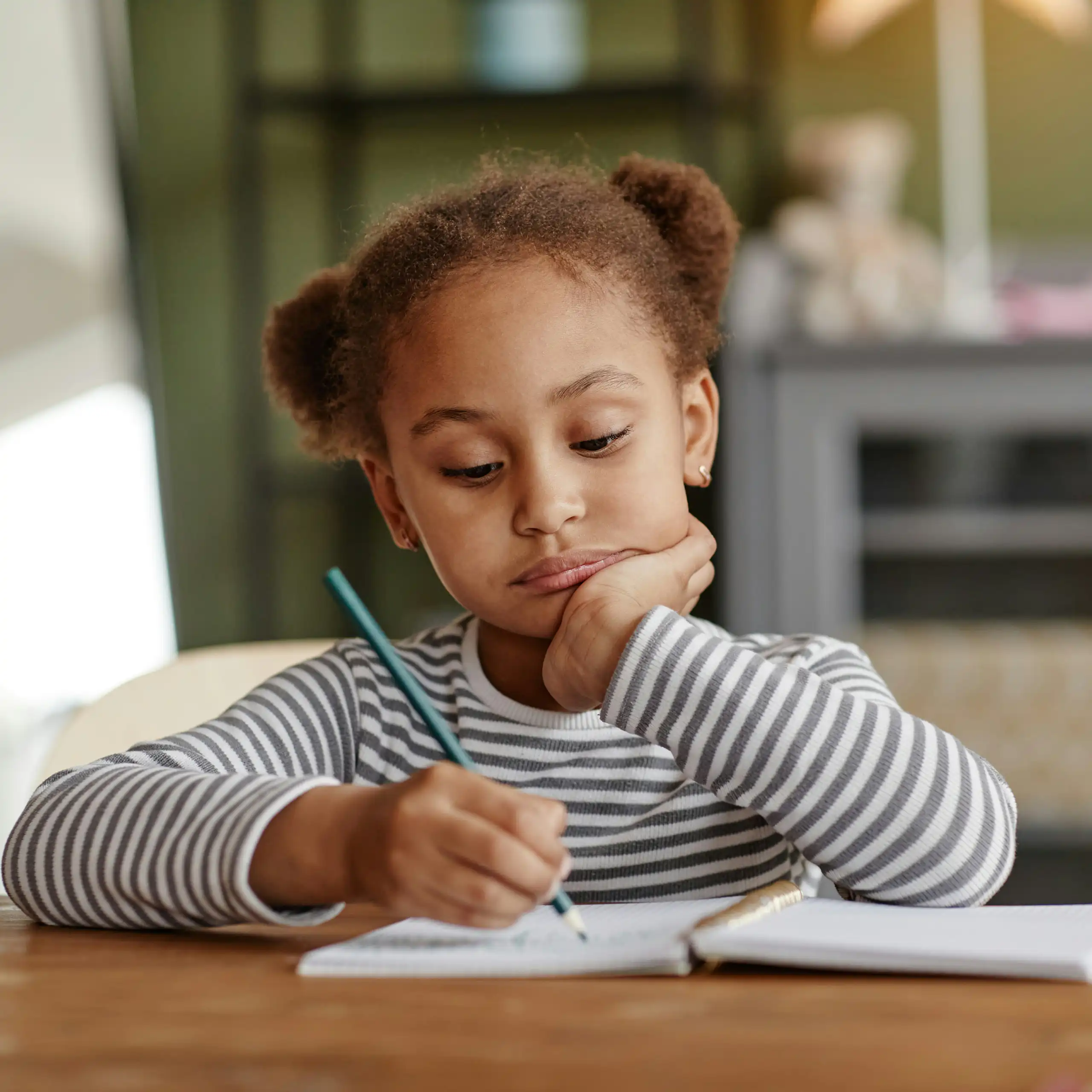 A girl in a striped tee sitting at a table with a notebook, resting her chin in her hand