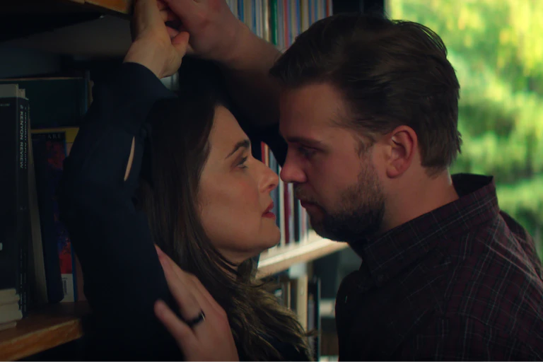 a man presses a woman against a bookshelf and they look at each other with longing