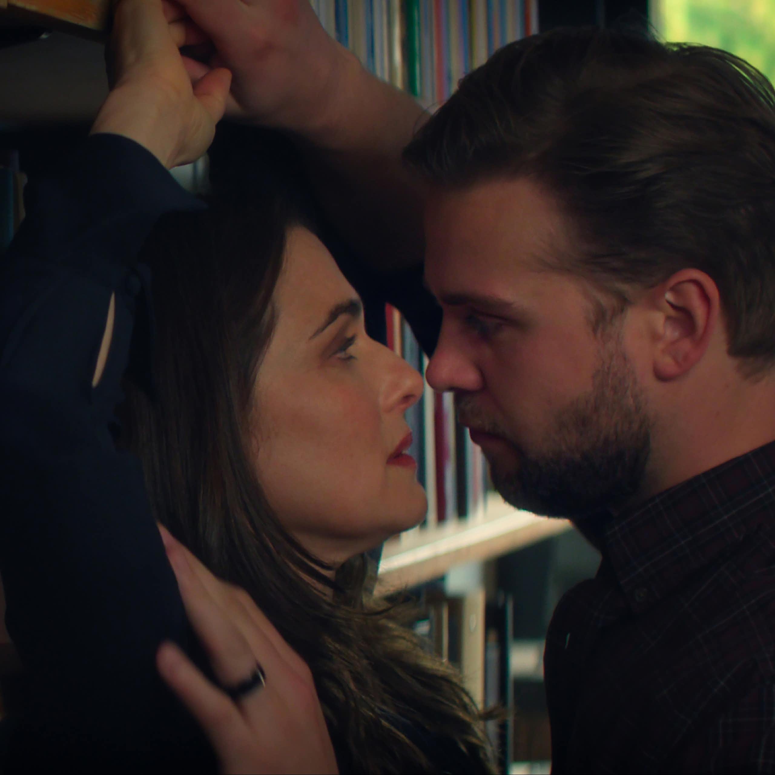 a man presses a woman against a bookshelf and they look at each other with longing