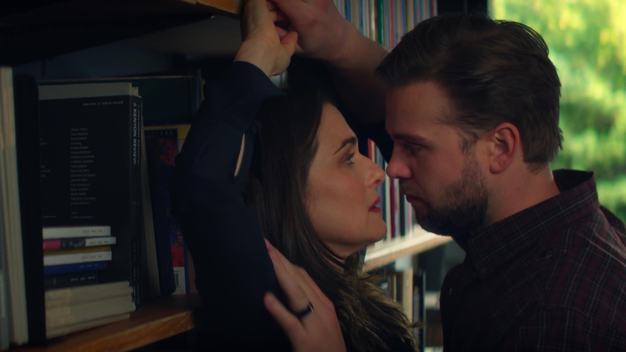 a man presses a woman against a bookshelf and they look at each other with longing