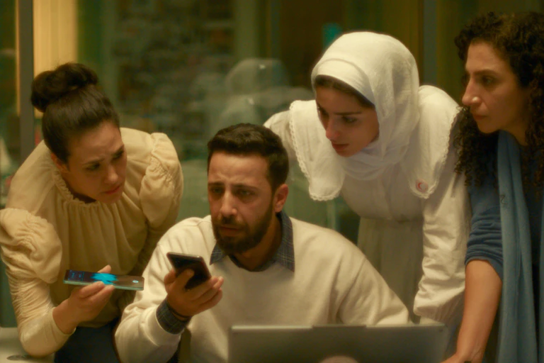 Three women, one in a headscarf, stand anxiously above a man seated with a phone in his hand.