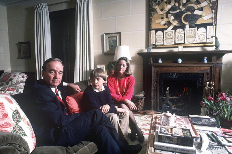 A man in a suit on a couch with a small boy, and a blonde woman in a red jumper