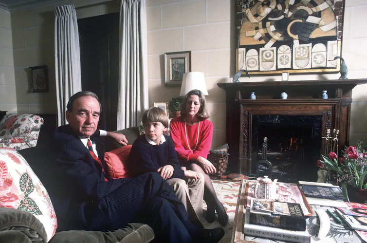 A man in a suit on a couch with a small boy, and a blonde woman in a red jumper
