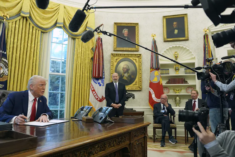 Donald Trump in Oval Office surrounded by cameras, Rupert Murdoch in background, in suit and sneakers