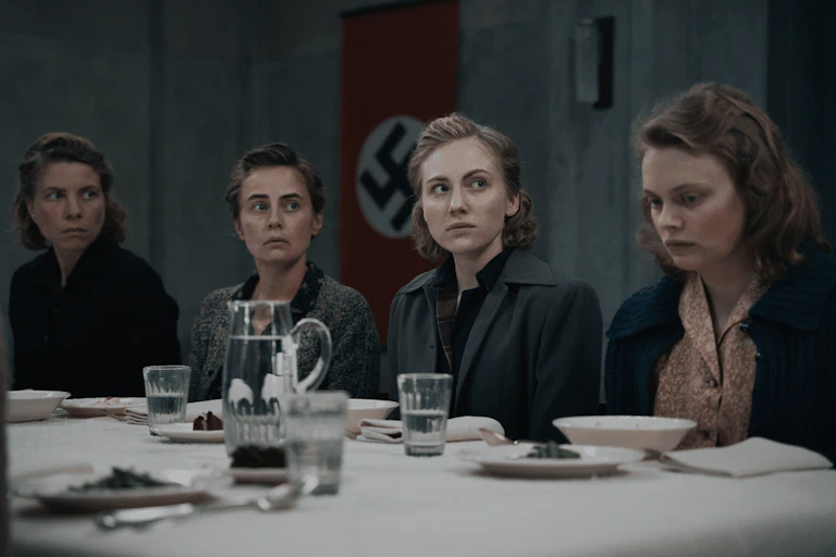 Four young women looking sombre seated at a table with the Nazi flag behind them.