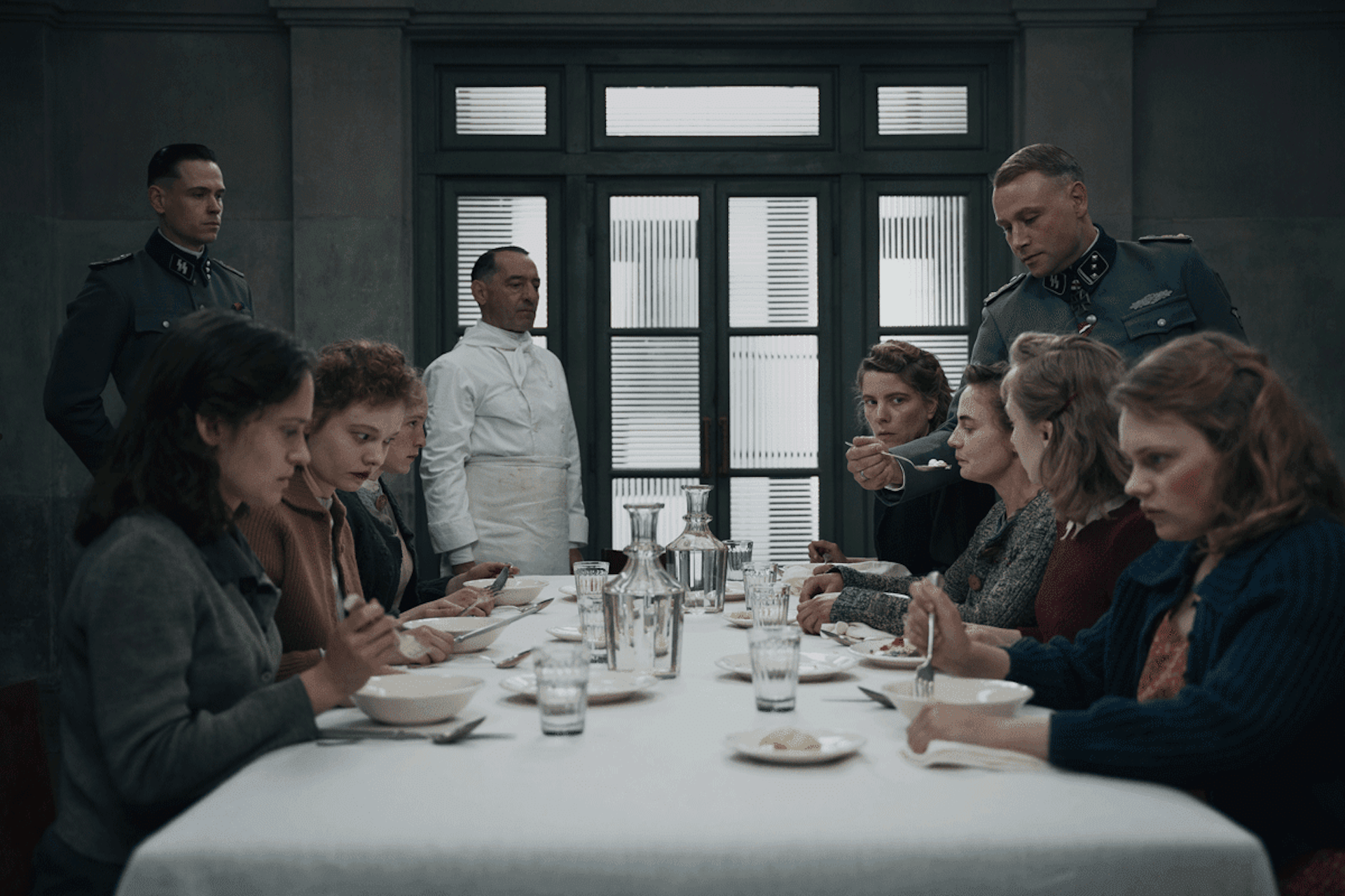 A chef, and two Nazi soldiers watch a group of women at a table tasting food.