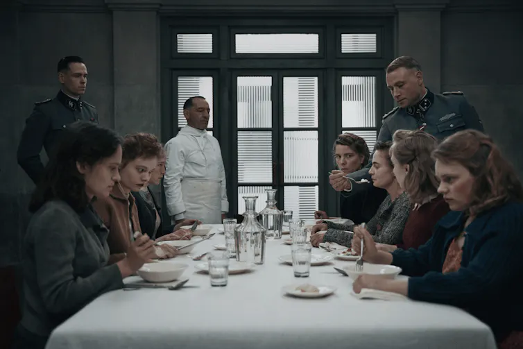 A chef, and two Nazi soldiers watch a group of women at a table tasting food.