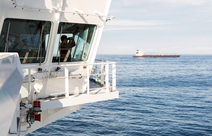a man inside the bridge of a large ship at sea looks through binoculars with another ship in the background