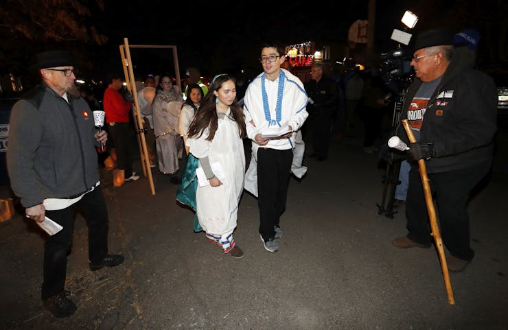 A young man and woman, both of whom wear white costumes, walk at the front of a small procession outside at night.