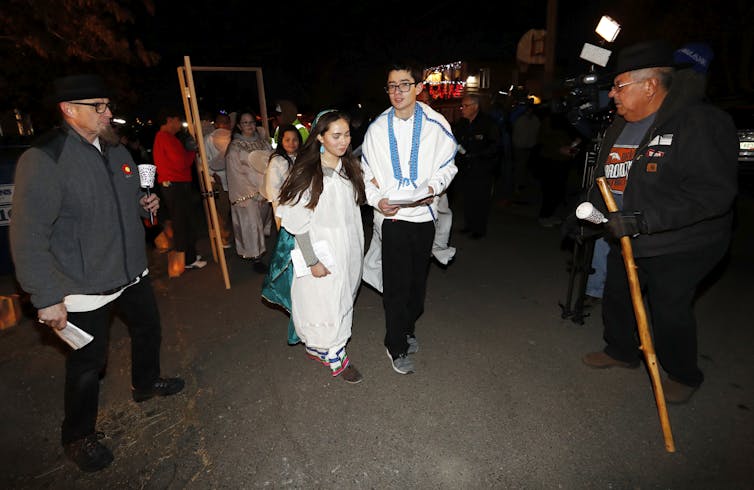 Un joven y una mujer, ambos vestidos con trajes blancos, caminan delante de una pequeña procesión afuera por la noche.