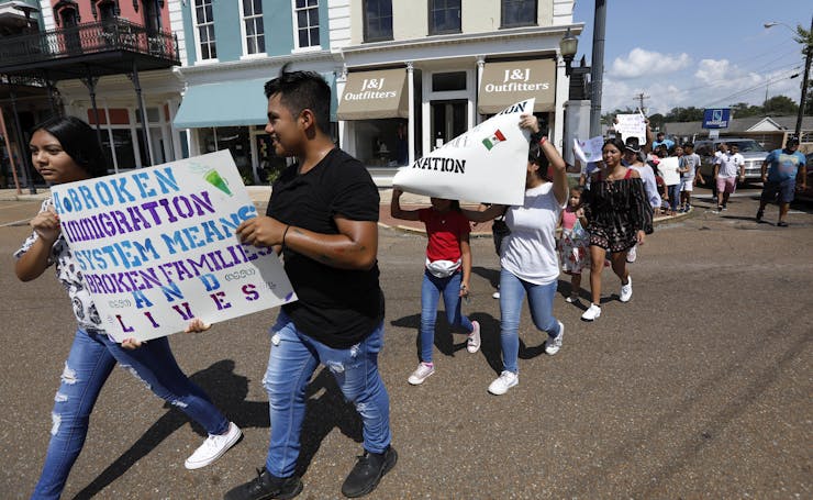A small group of boys and girls walk two-by-two through a town square, holding protest signs.