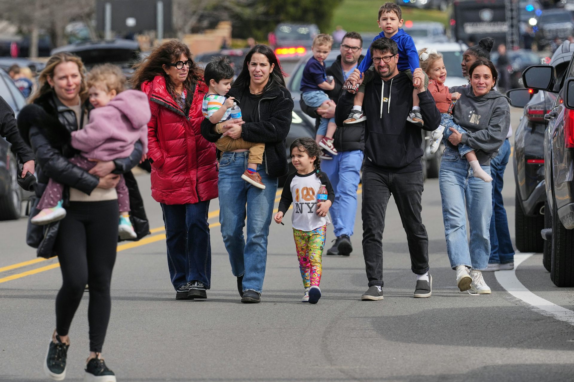 A small group of men and women in winter coats escort children down a paved road, with lots of cars in the background.