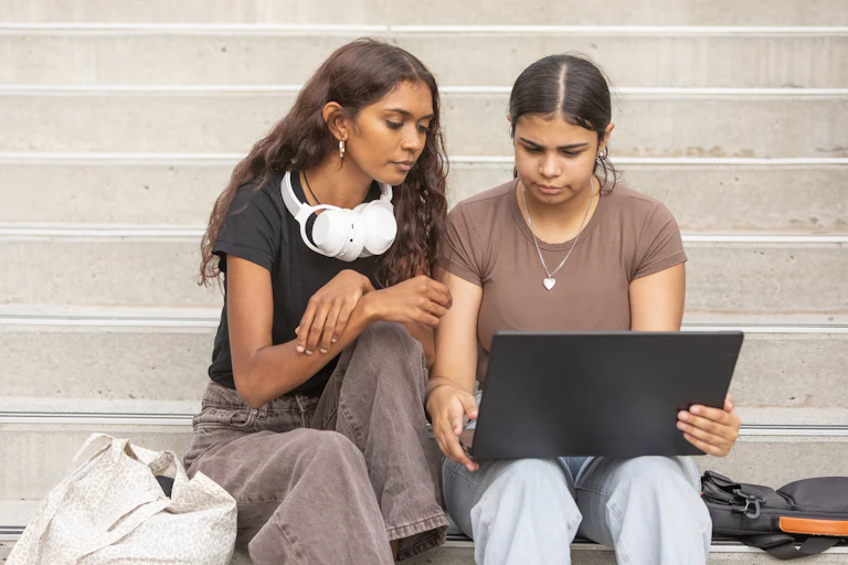 Two young women sit on a staircase and look at a laptop.
