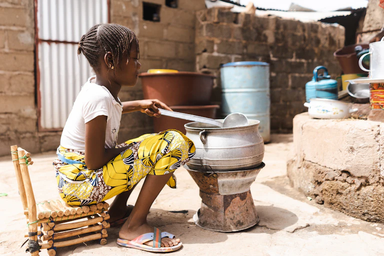 little African girl sitting stirring a pot on a stove.