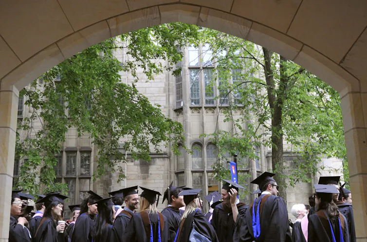 A small crowd of young people in black robes and flat black hats wait under a stone archway.